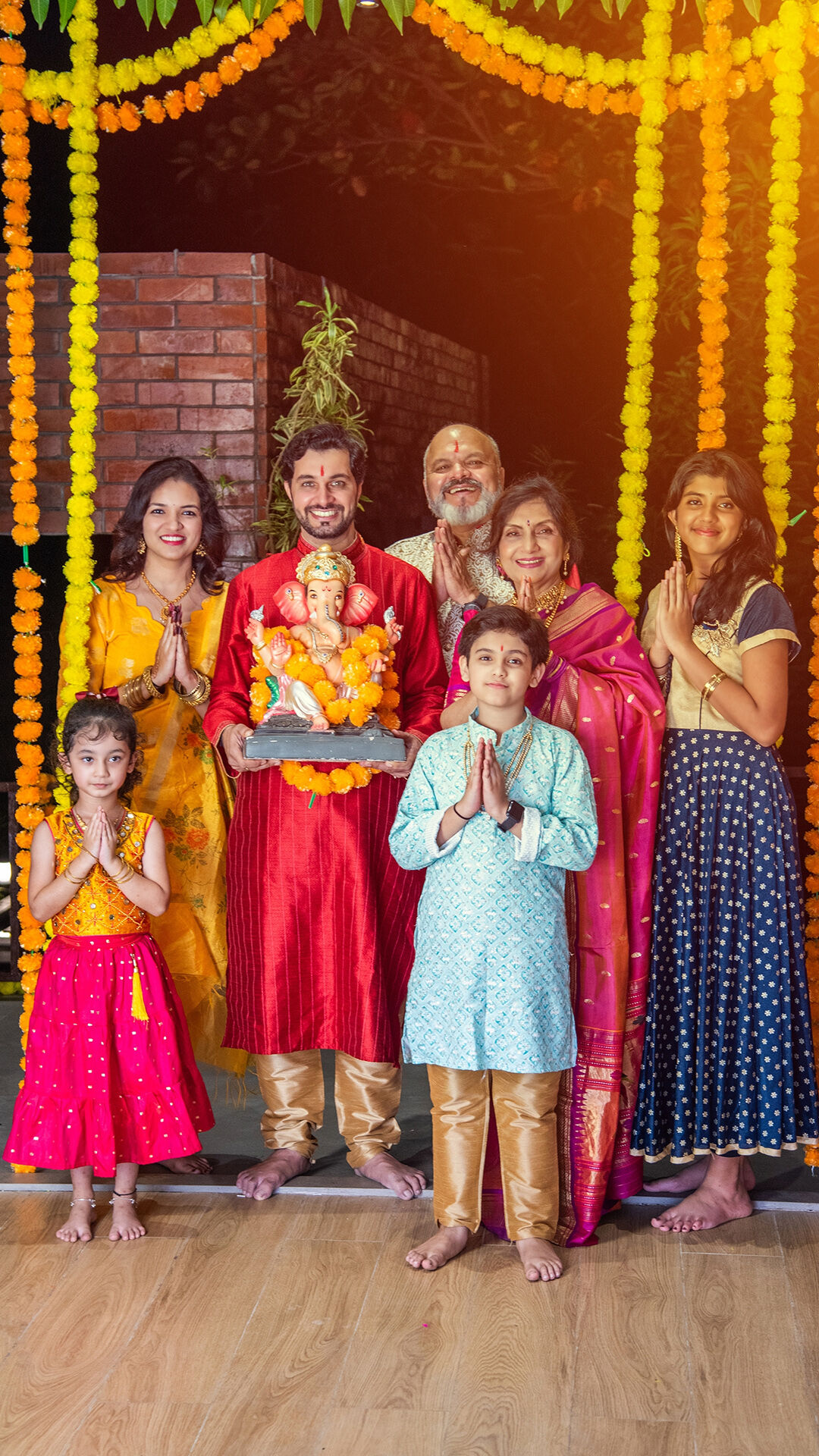 Smiling Indian family dressed in traditional attire posing with Lord Ganesha idol during Ganesh Chaturthi celebrations, surrounded by marigold flower decorations