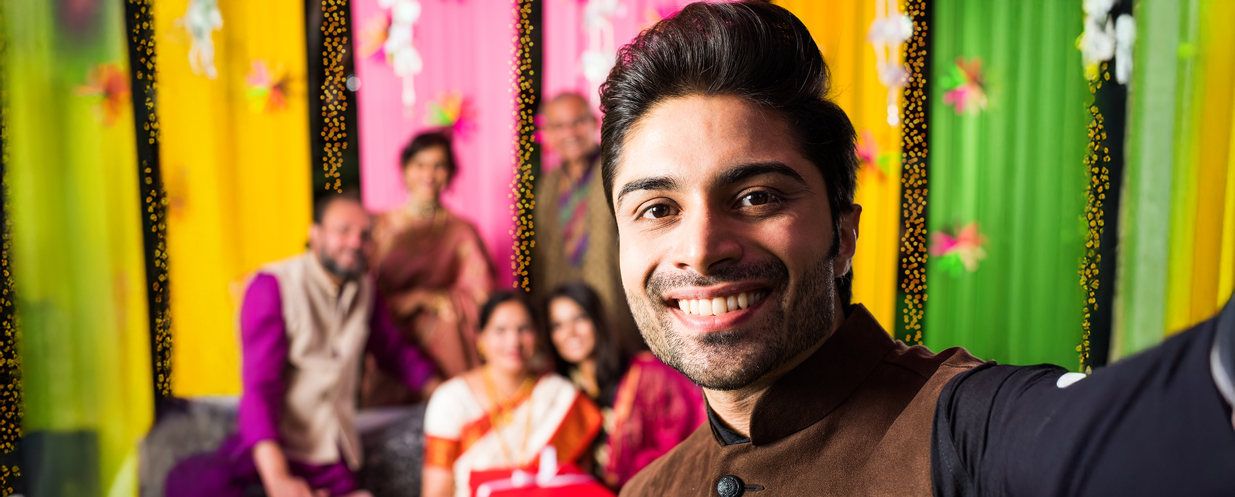 Smiling groom taking a selfie with family at a colorful Indian wedding selfie point decorated with drapes and fairy lights. Smiling groom taking a selfie with family at a colorful Indian wedding selfie point decorated with drapes and fairy lights.