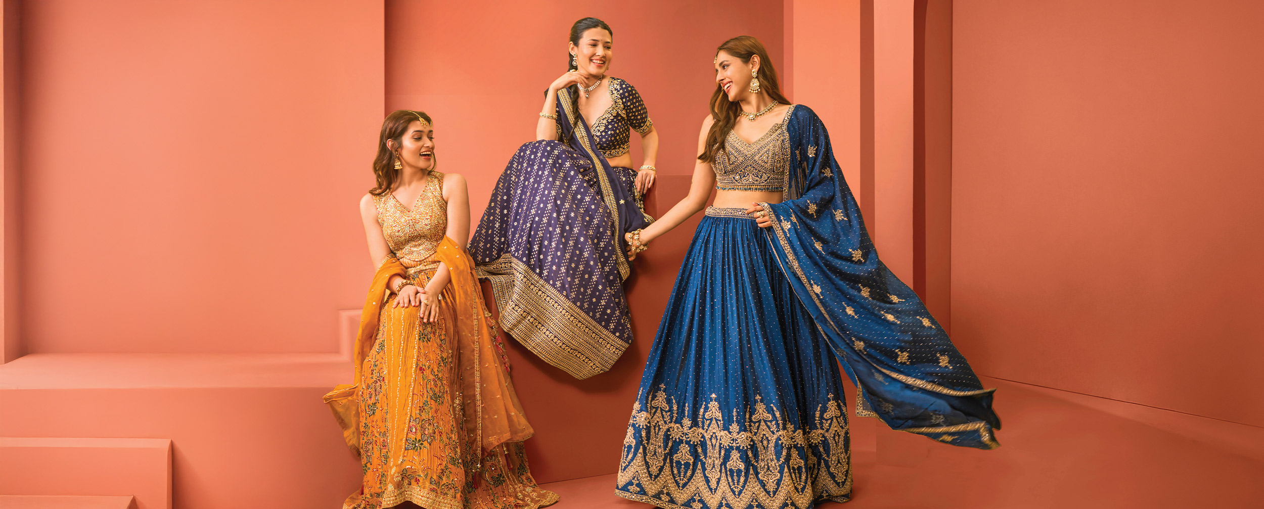Three women wearing vibrant lehengas in mustard, navy blue, and royal blue tones, showcasing trendy wedding guest outfit color choices against a coral backdrop