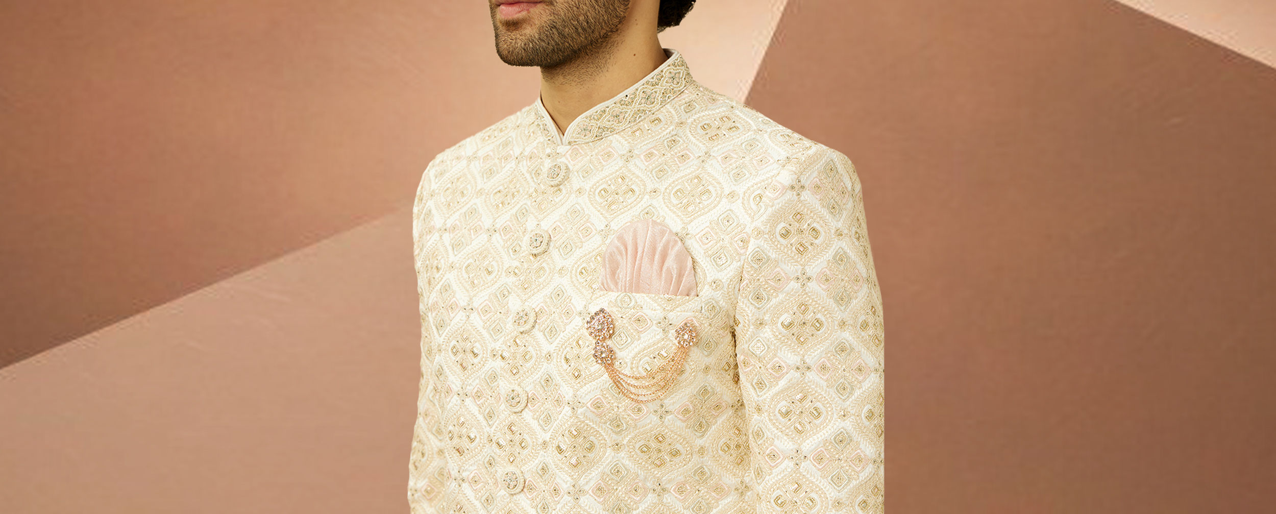 Ivory embroidered Sherwani jacket with a pink pocket square and rose gold brooch detail. Ivory embroidered Sherwani jacket with a pink pocket square and rose gold brooch detail.