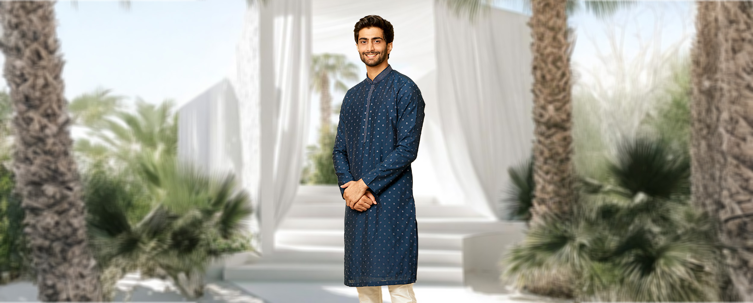 Man in a dark blue patterned kurta with churidar pajamas, posing against a white floral backdrop.