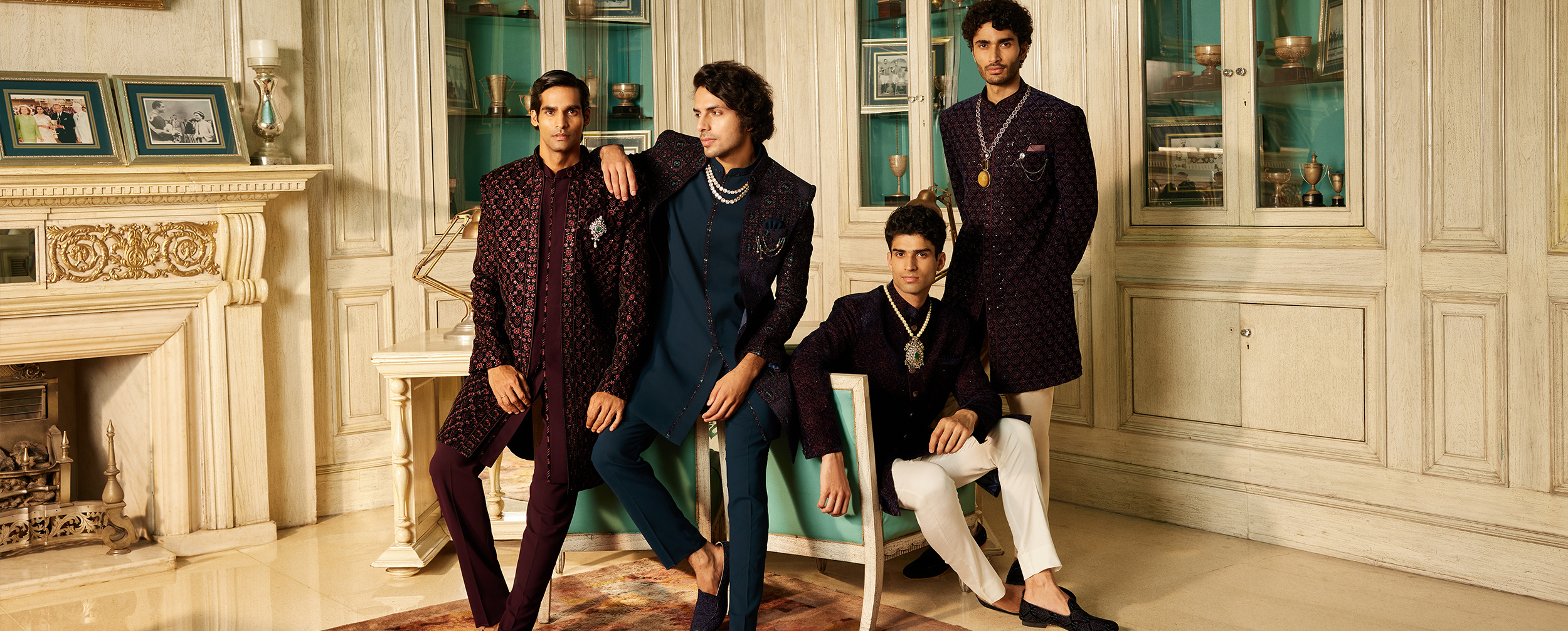 Four men in regal Indo-western wedding outfits featuring embroidered jackets, layered necklaces, brooches, and mojaris Four men in regal Indo-western wedding outfits featuring embroidered jackets, layered necklaces, brooches, and mojaris