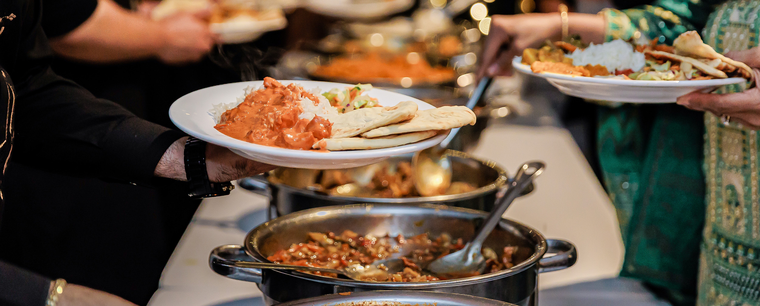  Indian wedding buffet with hands serving butter chicken (curry) and rice onto a white plate.