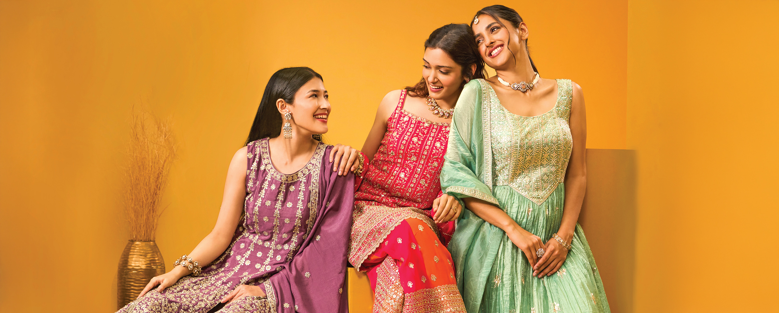 Three women wearing colorful embellished Indian ethnic outfits perfect for sangeet or wedding functions, posing against a mustard backdrop