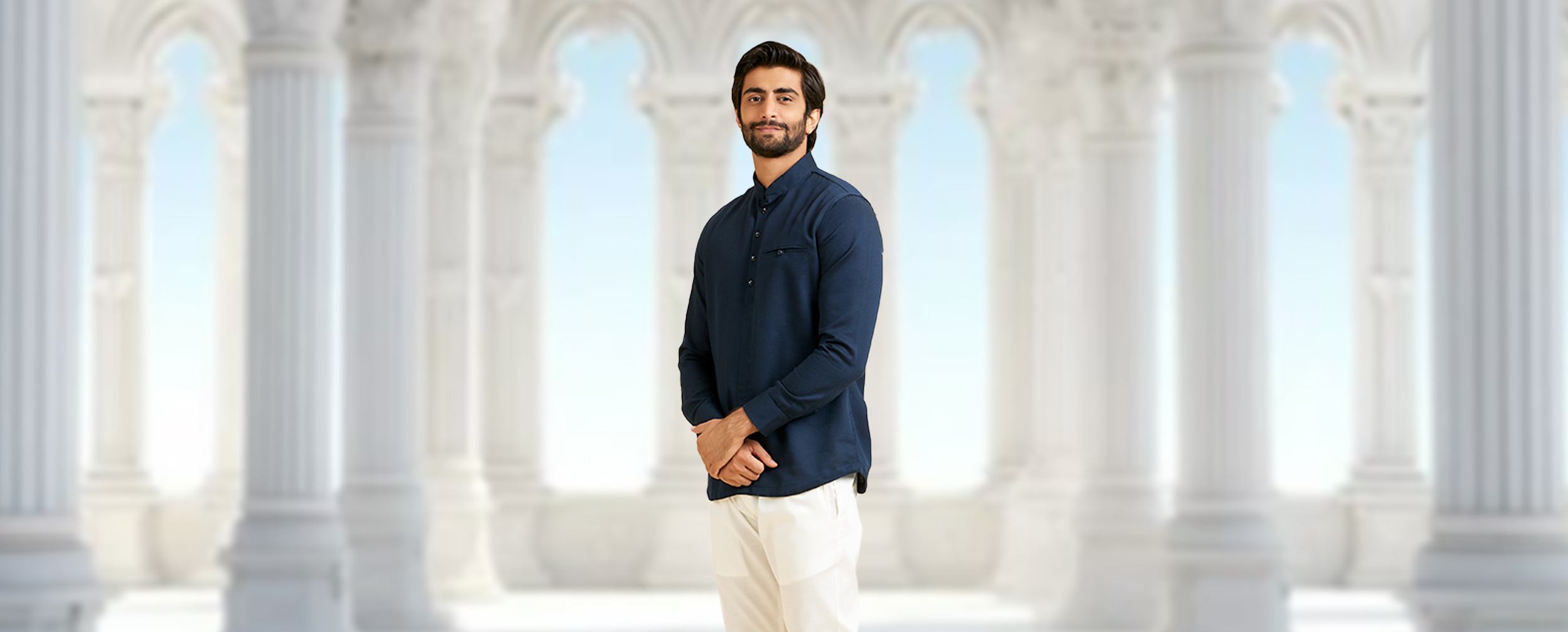  Man in a navy blue long-sleeved shirt kurta and white trousers, posing in front of white arches.