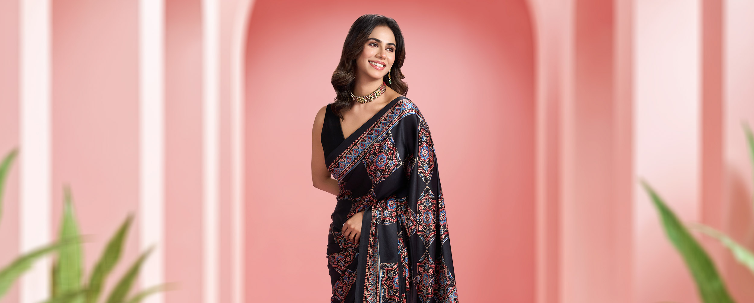 Woman in a black printed saree with traditional motifs, styled with sleeveless blouse and choker necklace against a pink background.