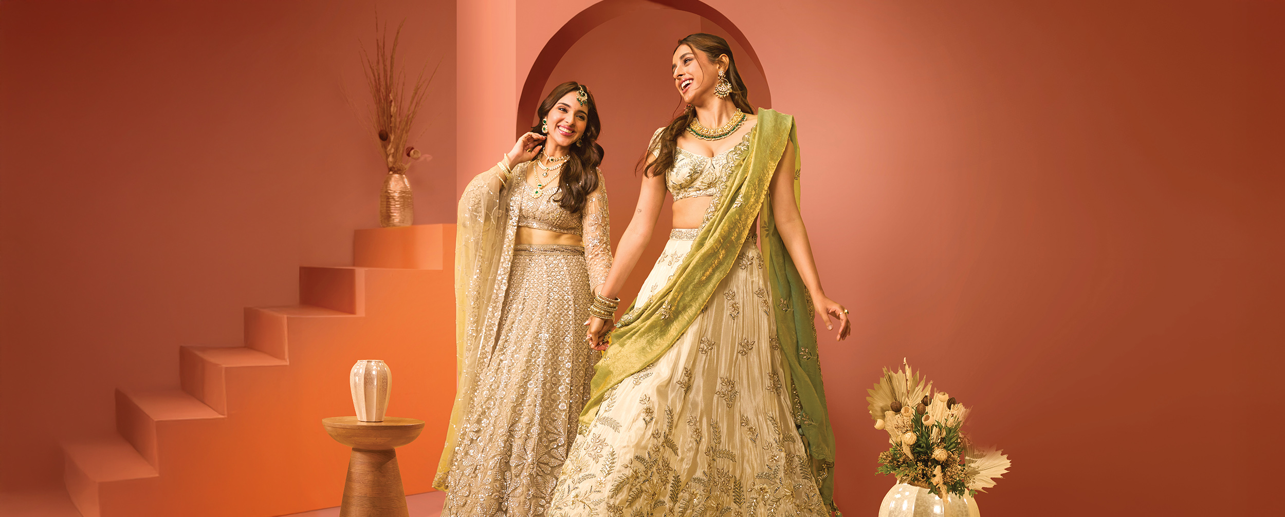 Two women in embellished designer lehengas, styled with statement jewelry and contrasting dupattas, posing joyfully against a peach backdrop. Two women in embellished designer lehengas, styled with statement jewelry and contrasting dupattas, posing joyfully against a peach backdrop.