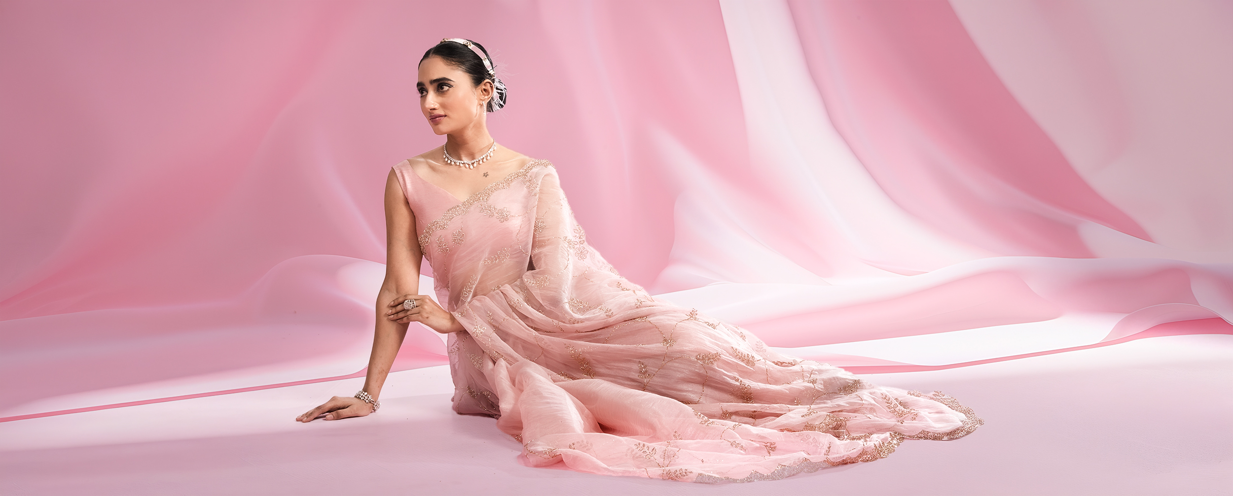 Elegant woman in a pastel pink embroidered saree, styled with a sleek bun and delicate jewelry, posing gracefully against a soft draped pink background