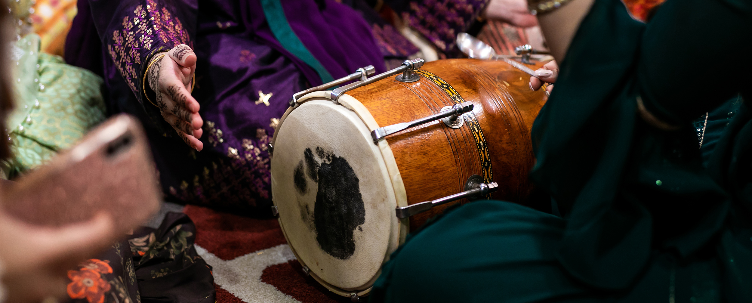 Close-up of a traditional Indian dhol being played during a vibrant pre-wedding celebration, surrounded by women in festive attire. Close-up of a traditional Indian dhol being played during a vibrant pre-wedding celebration, surrounded by women in festive attire.