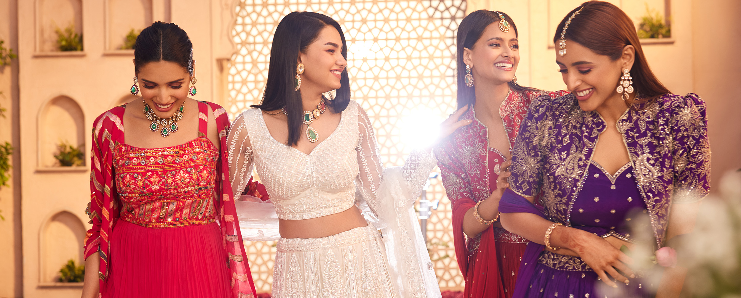 Four women in vibrant wedding guest attire, including a white lehenga, a pink anarkali, a purple lehenga, and a red outfit. Four women in vibrant wedding guest attire, including a white lehenga, a pink anarkali, a purple lehenga, and a red outfit.