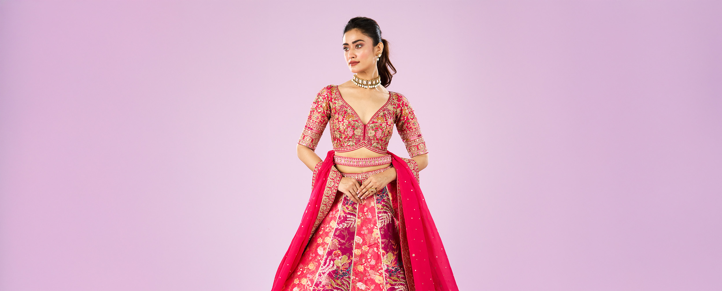 Bride in a vibrant pink embroidered bridal blouse and lehenga with floral motifs, styled with a statement choker necklace and matching dupatta against a pastel pink background.