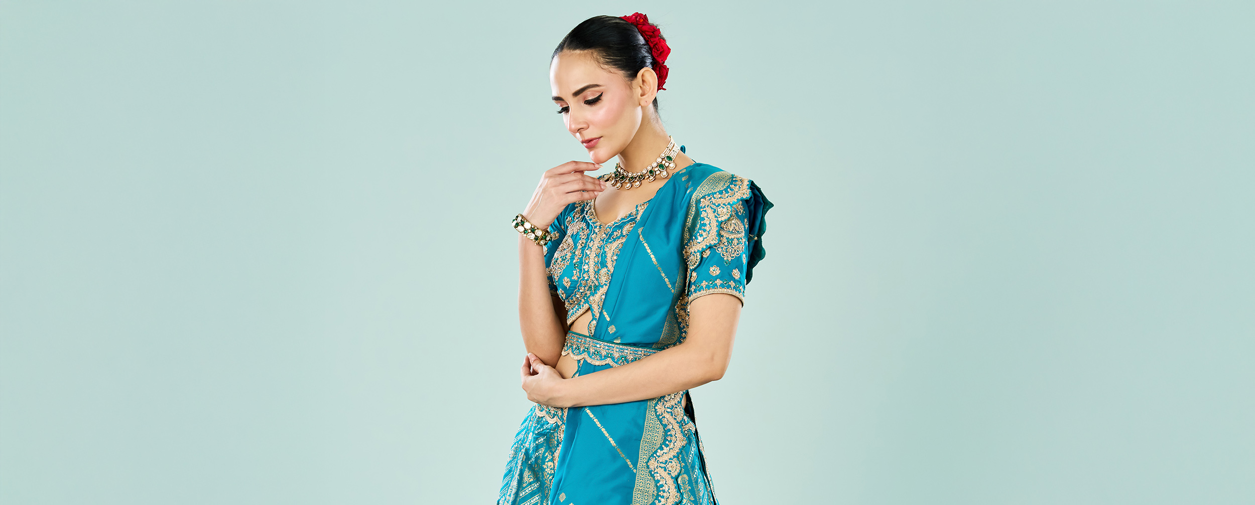 Bride in royal blue embroidered lehenga with gold zari work, styled with traditional choker necklace and red floral hair bun for modern bridal look