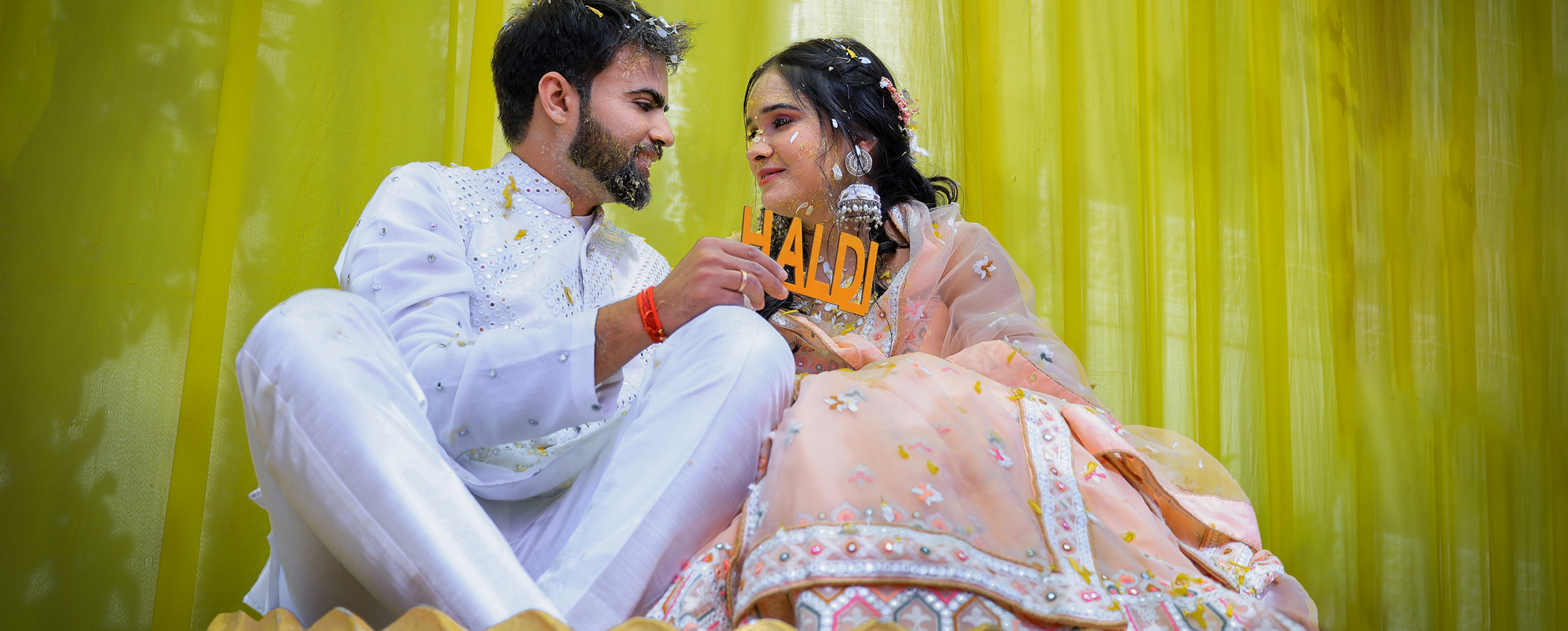 Bride and groom joyfully holding a 'Haldi' sign during their vibrant pre-wedding Haldi ceremony. Bride and groom joyfully holding a 'Haldi' sign during their vibrant pre-wedding Haldi ceremony.