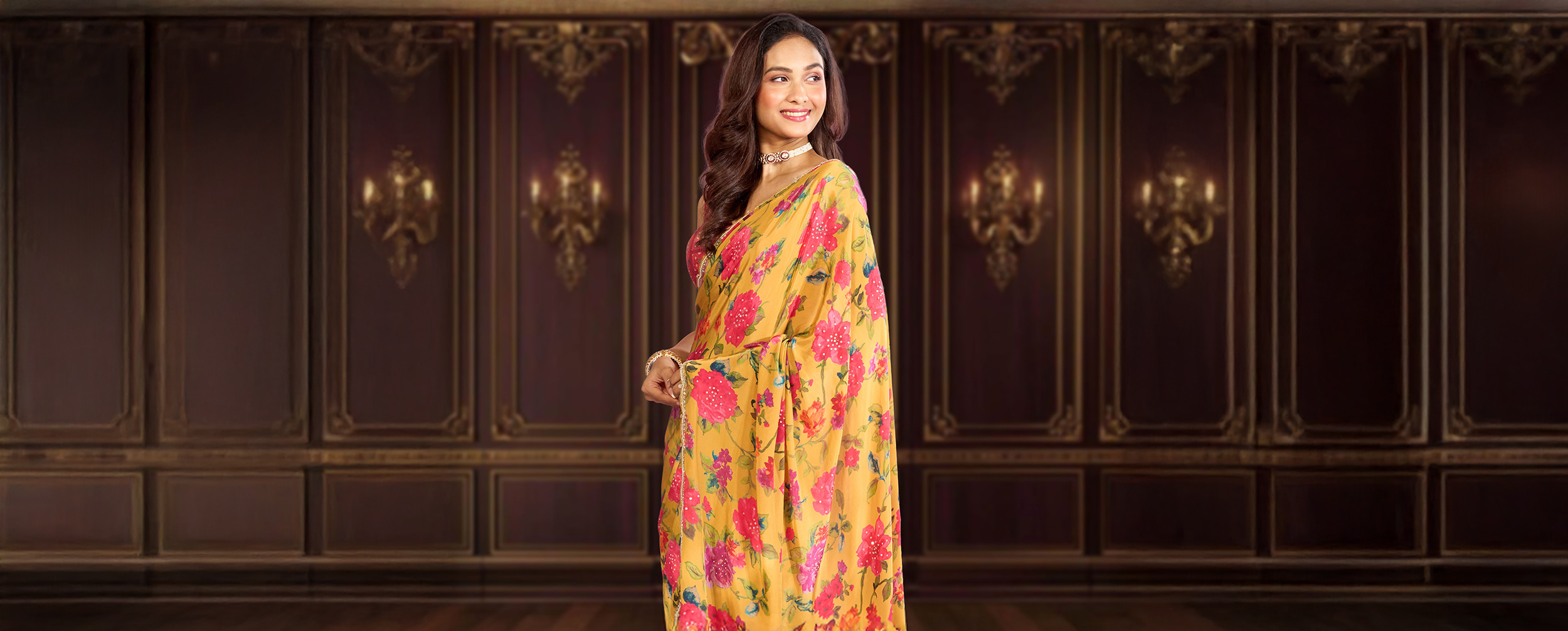 Smiling woman in a bright yellow saree with vibrant pink floral prints, styled with a choker necklace, standing against a royal backdrop, representing Navratri festive fashion.