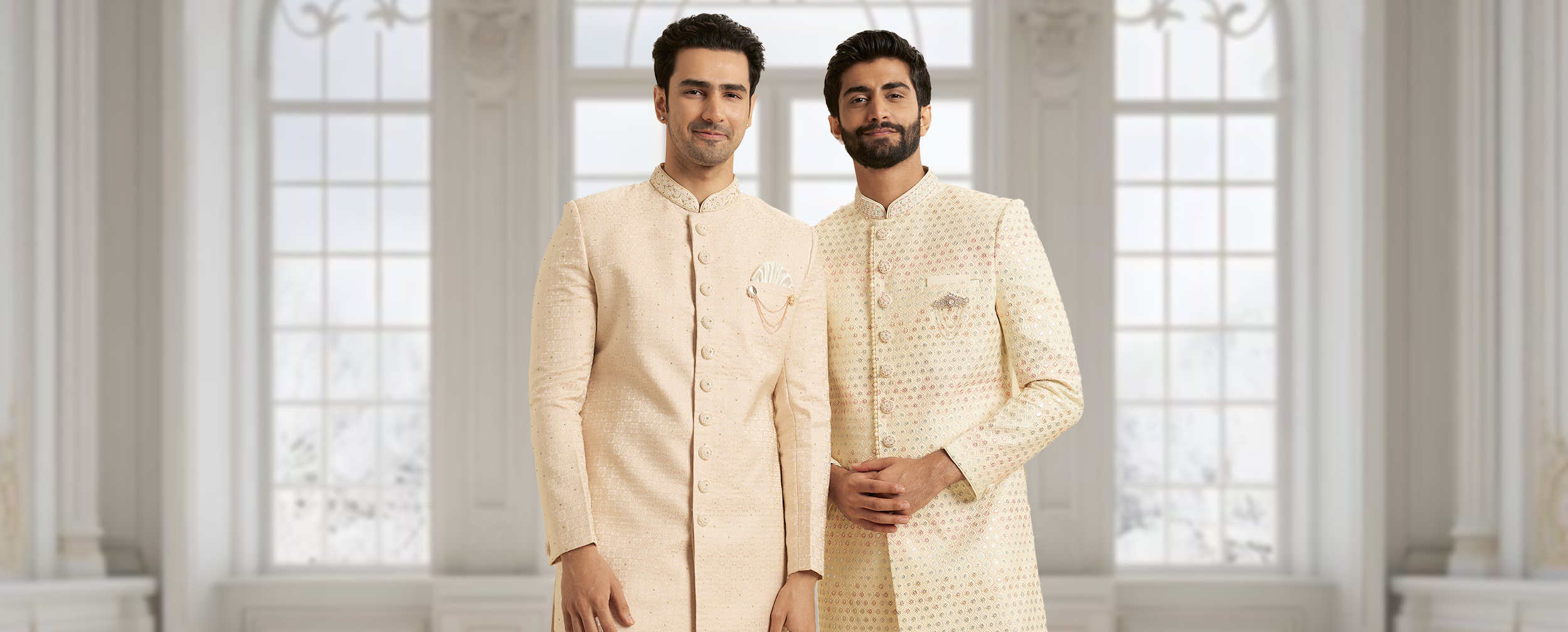 Two men wearing elegant cream and beige sherwanis with intricate embroidery, posing in a bright royal-inspired interior.