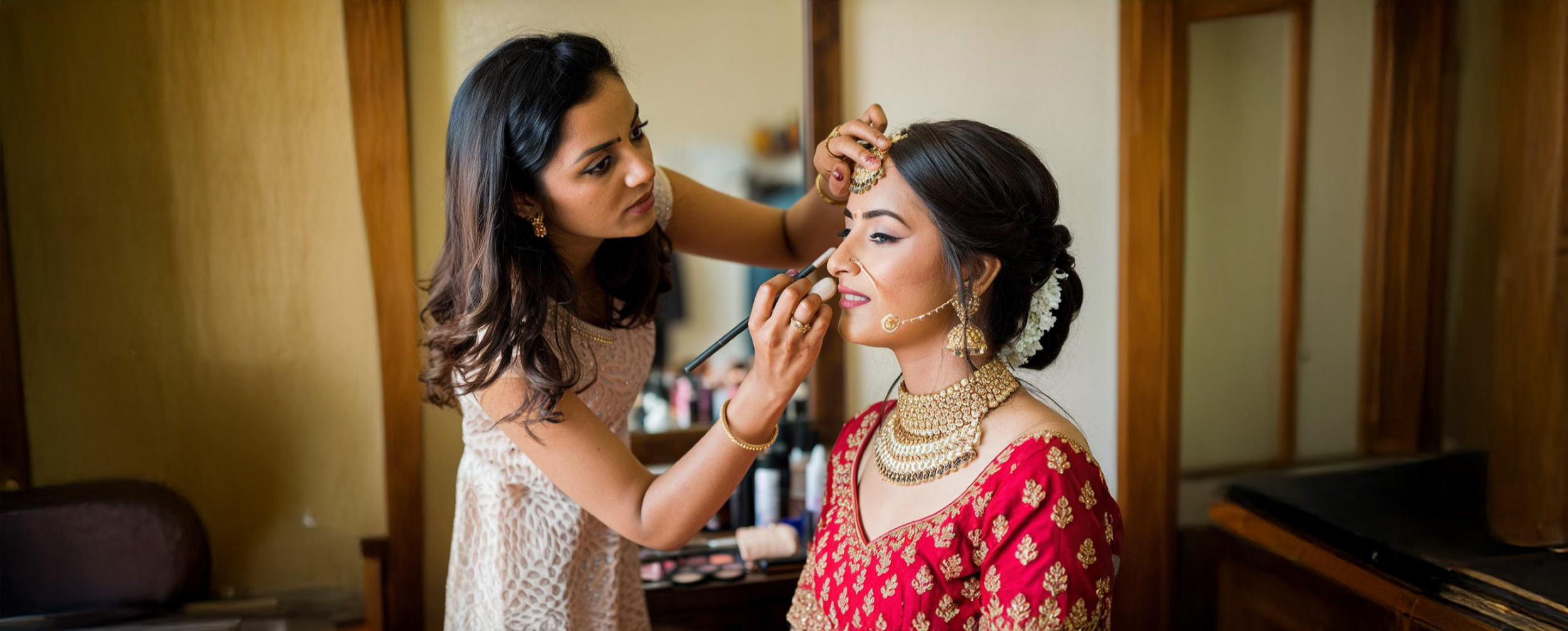 Makeup artist applying eye makeup to an Indian bride dressed in a red and gold bridal outfit, adorned with traditional jewelry and floral hair accessories Makeup artist applying eye makeup to an Indian bride dressed in a red and gold bridal outfit, adorned with traditional jewelry and floral hair accessories