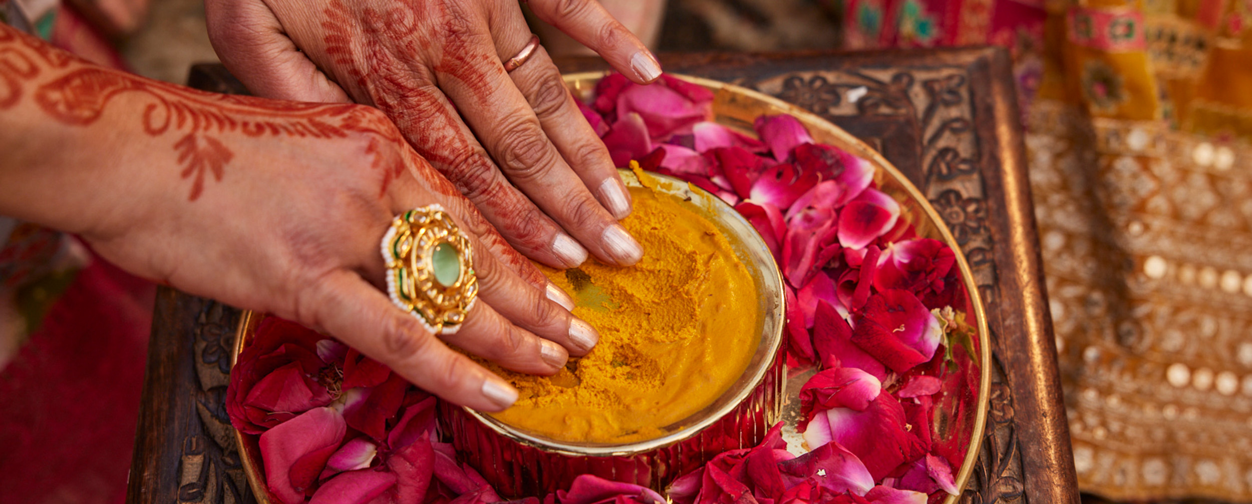 Hands adorned with mehndi and rings dipping into haldi paste surrounded by rose petals, symbolizing the Haldi ceremony in a Hindu wedding ritual.
